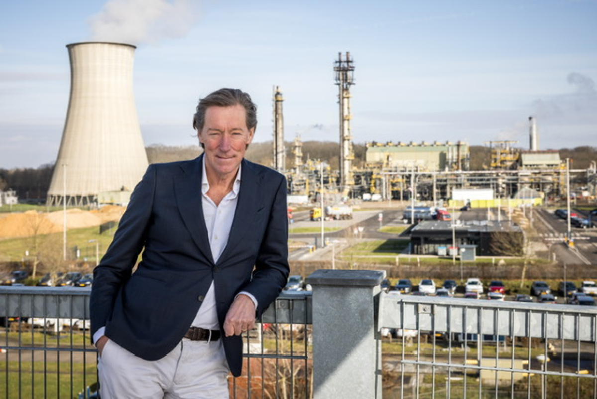 Een man in een donkerblazer staat op een balustrade, met een industriegebied op de achtergrond.