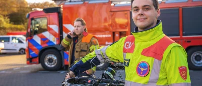 Twee leden van de brandweer, één met drone, staan voor een brandweerwagen.