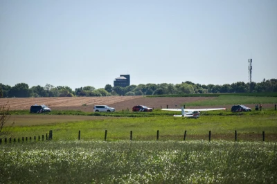 Een sportvliegtuigje staat stil aan het einde van een landweg met auto's van de hulpdiensten ernaast en groene velden op de achtergrond.