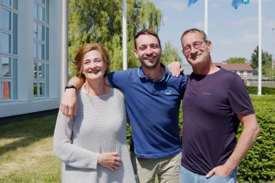 Drie mensen staan samen buiten, lachend, met een groen landschap en blauwe lucht achter hen.