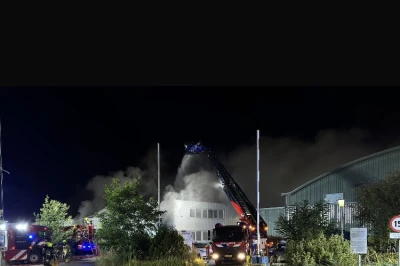 Brand op een industrieel terrein, met brandweermensen en rookpluimen in de lucht.
