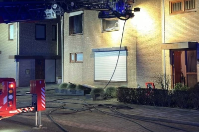 Brandweerman op ladder bij een brand in een huis, met rook en een brandslang op de grond.