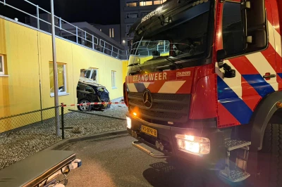 Brandweerwagen voor een gele muur, met een ongeval in de achtergrond bij nacht.