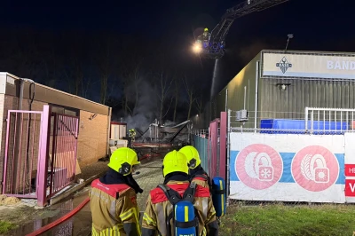 Brandweerlieden in gele helmen kijken naar een brand bij een gebouw in de nacht.