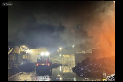Rook en vlammen in een afgebrande loods met een brandweertruck in de verte.