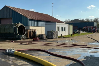Een industrieel terrein met een gebouw, brandslangen en waterplassen in de zon.
