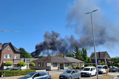 Zwart rook stijgt op achter huizen onder een blauwe lucht. Autos op de voorgrond.