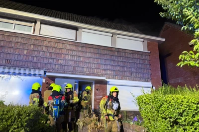 Brandweerlieden in uniform bij een woning met rook en een nachtelijke achtergrond.