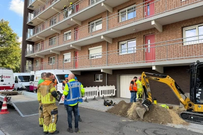 Hulpdiensten en bouwvakkers rond een graafmachine bij een appartementengebouw.