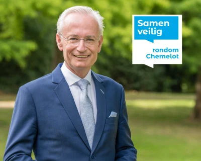 Burgemeester Sittard-Geleen in blauw pak staat glimlachend in een groene parkachtige omgeving.