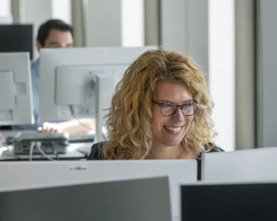 Een vrouw met krullend haar en een bril glimlacht terwijl ze naar een computer kijkt.