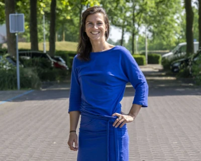 Vrouw in een blauwe outfit poseert lachend op een parkeerplaats, omringd door bomen.